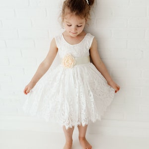 Pode incluir: Uma menina pequena vestindo um vestido branco de renda com uma faixa de cetim e um detalhe de flor de p&ecirc;ssego. O vestido tem um decote redondo e &eacute; sem mangas. A menina est&aacute; em p&eacute; em um fundo branco.