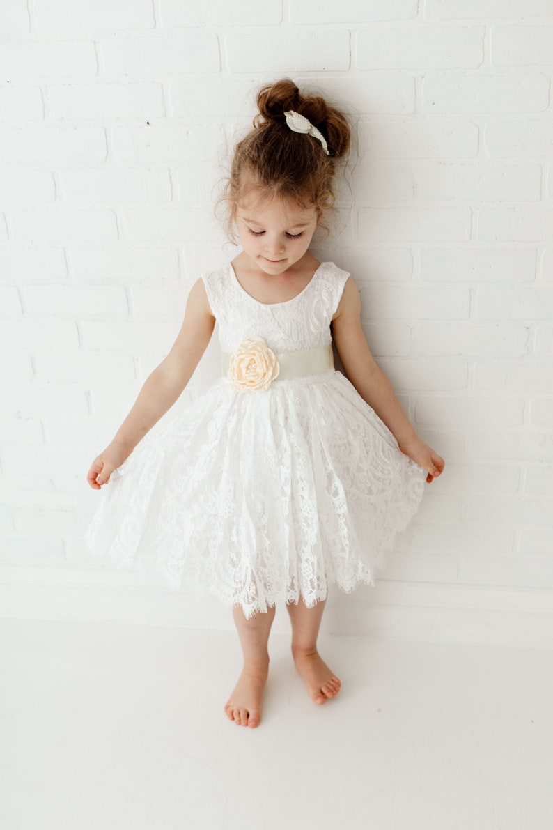 Pode incluir: Uma menina pequena vestindo um vestido branco de renda com uma faixa de cetim e um detalhe de flor de p&ecirc;ssego. O vestido tem um decote redondo e &eacute; sem mangas. A menina est&aacute; em p&eacute; em um fundo branco.