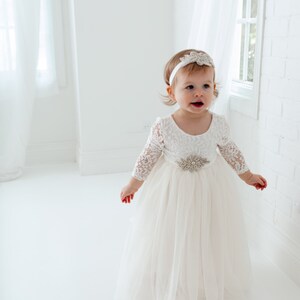 Pu&ograve; includere: Una bambina che indossa un abito bianco di tulle con maniche di pizzo e una cintura con gioielli. Indossa una fascia bianca con un gioiello.