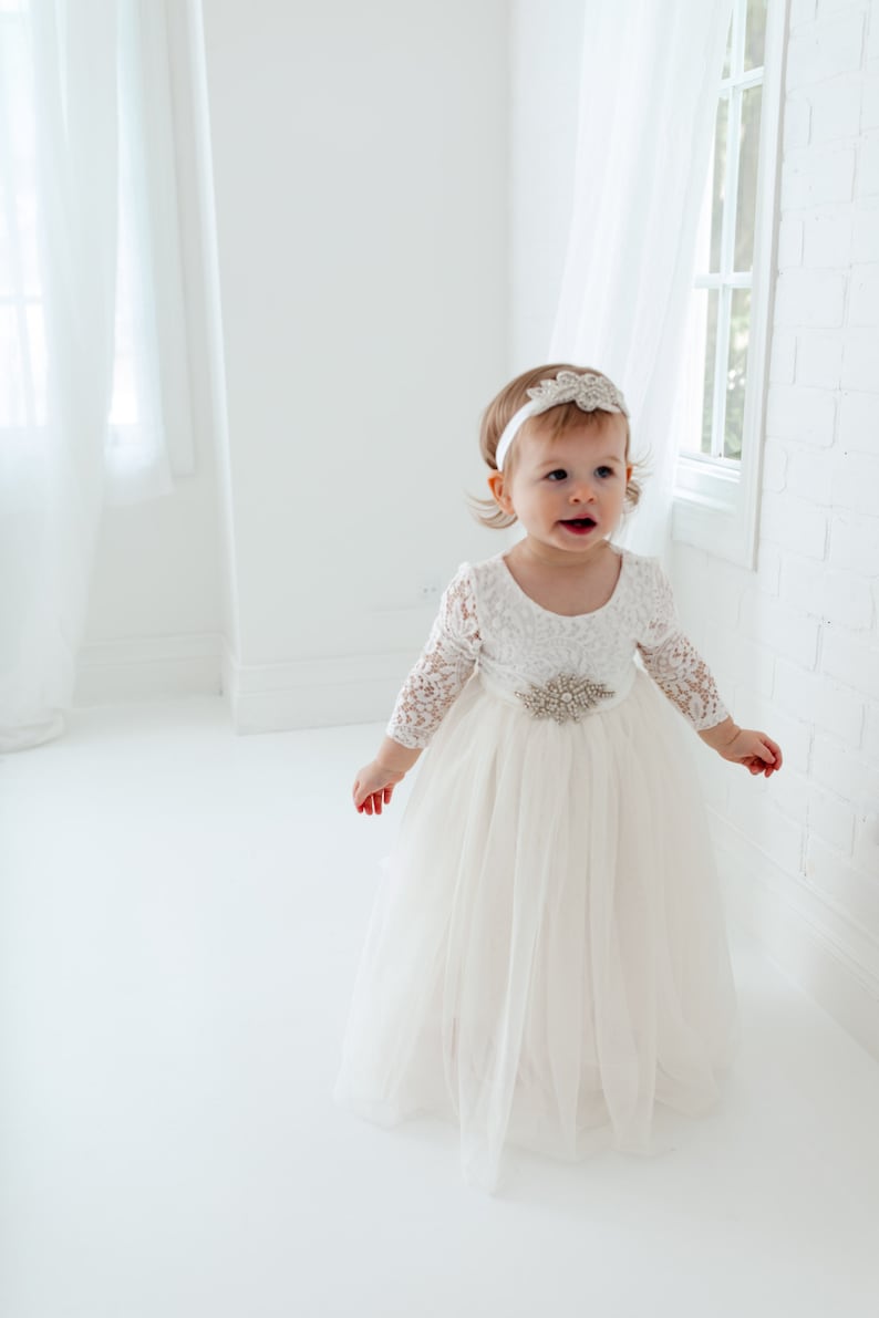 Pu&ograve; includere: Una bambina che indossa un abito bianco di tulle con maniche di pizzo e una cintura con gioielli. Indossa una fascia bianca con un gioiello.