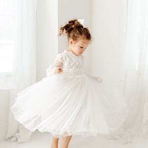 May include: A young girl wearing a white tulle dress with lace detailing. The dress has long sleeves and a full skirt. The girl is standing on a white background and is twirling the skirt of her dress.
