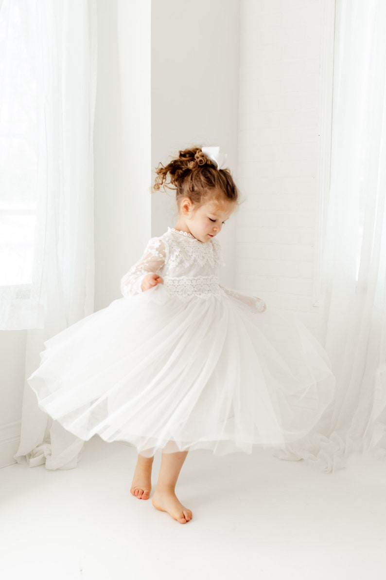 May include: A young girl wearing a white tulle dress with lace detailing. The dress has long sleeves and a full skirt. The girl is standing on a white background and is twirling the skirt of her dress.