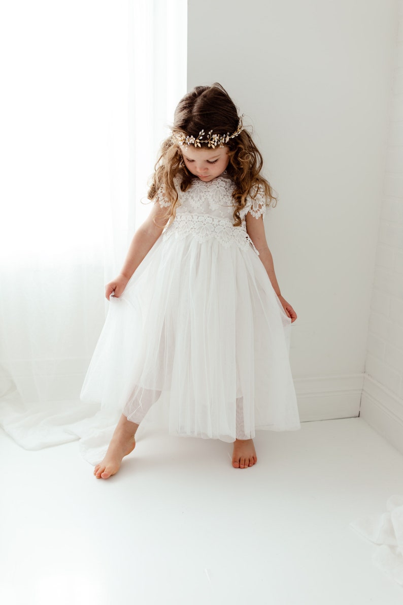 May include: A young girl in a white tulle dress with lace detailing and a gold floral crown. The dress has short sleeves and a flowing skirt. The girl is barefoot in a bright, white room.