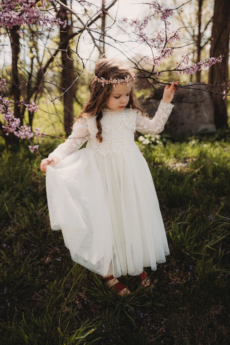 May include: A long-sleeved white dress featuring a lace bodice with a high neckline and a layered tulle skirt. The garment is paired with a delicate floral headpiece made of small pink and white blossoms, and brown sandals are visible at the base.