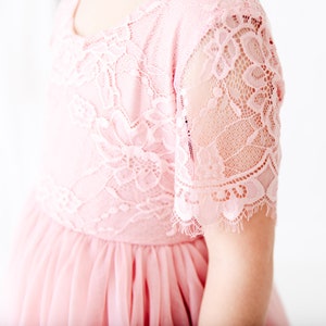 Pode incluir: Um vestido de tule rosa com um corpete de renda e mangas curtas. O vestido tem uma renda festonada.