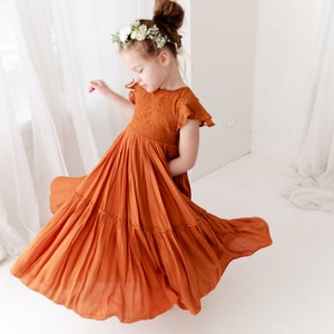 Op de afbeelding: Een jong meisje draagt een roestkleurige gelaagde jurk met een bloemenkrans. De jurk heeft een kantboord en korte mouwen. Het meisje draait in de jurk en de stof stroomt om haar heen.