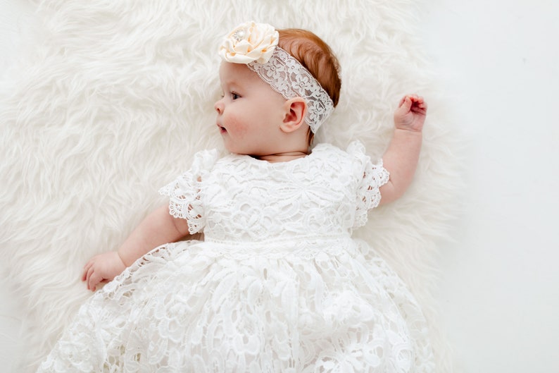 May include: A baby wearing a white lace dress with a flower headband. The baby is lying on a white fluffy surface.