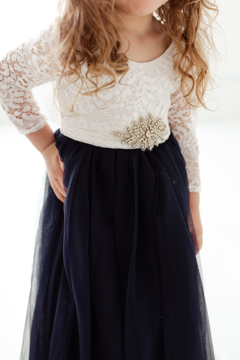 May include: A white lace dress with long sleeves and a navy blue tulle skirt. The dress has a white sash with a silver and crystal embellished brooch.