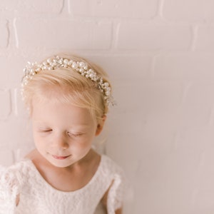 May include: A young girl with blonde hair wearing a white dress and a pearl and silver headband. She has her eyes closed and is looking down.