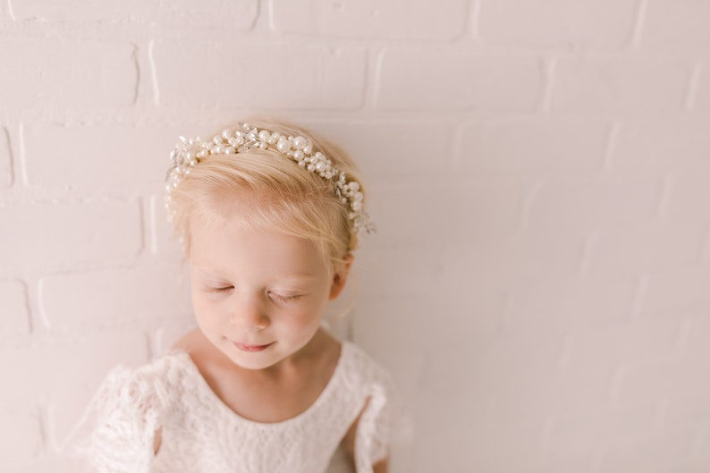 May include: A young girl with blonde hair wearing a white dress and a pearl and silver headband. She has her eyes closed and is looking down.