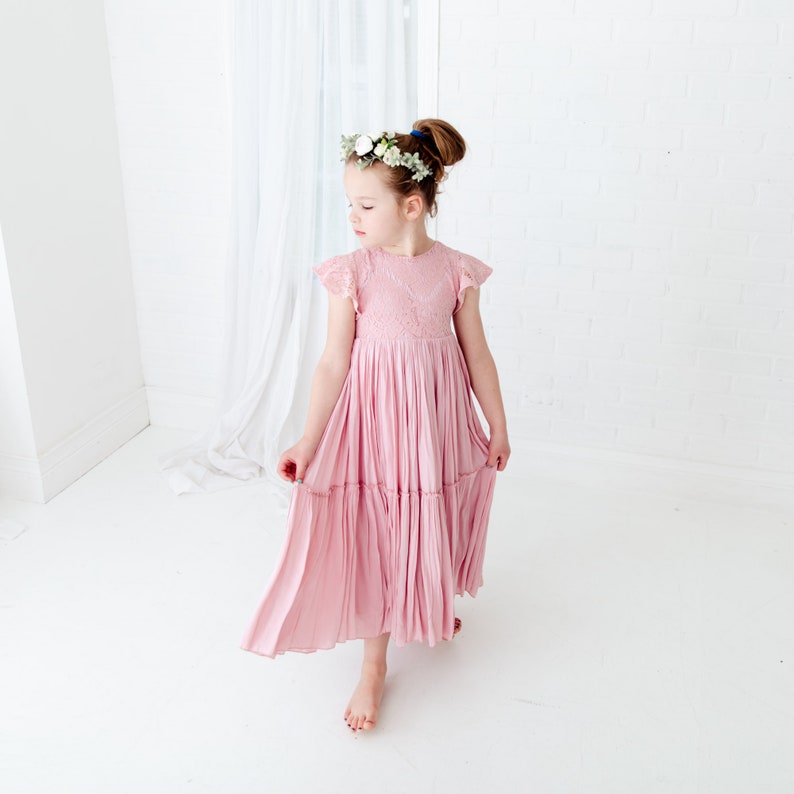 May include: A young girl wearing a pink, tiered, lace-trimmed dress with a floral crown. The dress has a gathered skirt and short sleeves.