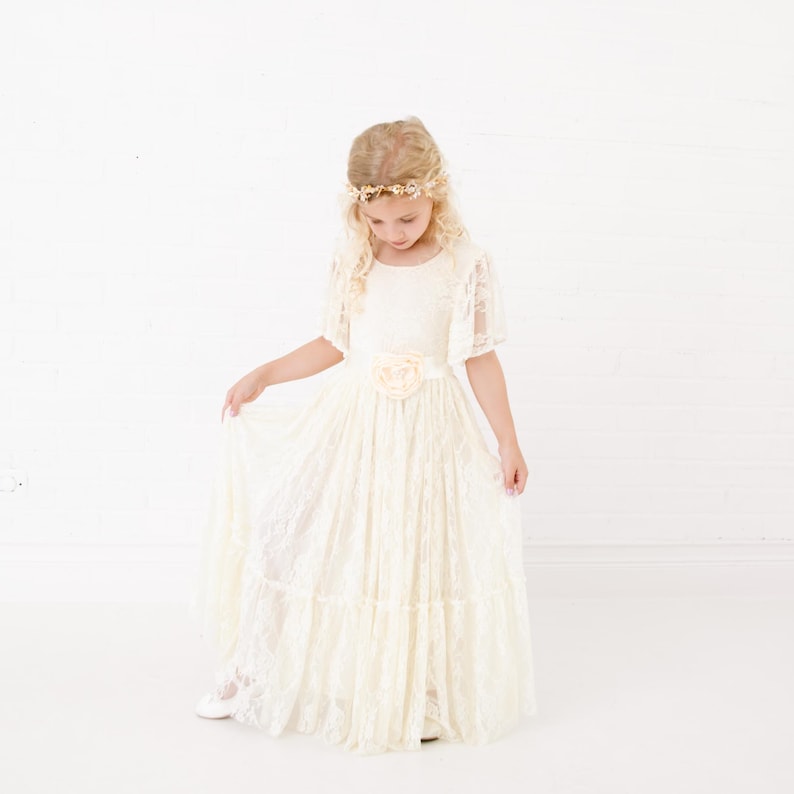Peut inclure: Une jeune fille portant une robe en dentelle blanche avec un accent floral &agrave; la taille. Elle porte une couronne de fleurs et tient la jupe de la robe.