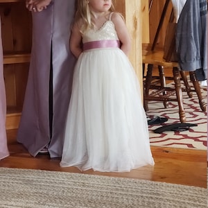 May include: A young girl wearing a white tulle dress with a pink sequined bodice and a pink satin sash stands next to a woman in a lavender satin dress. The girl is looking to the right and the woman is looking down.