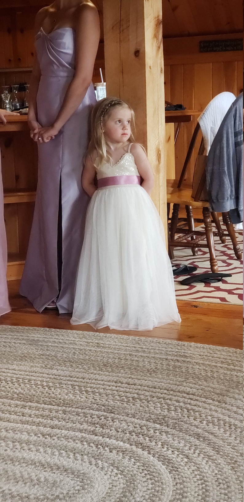 May include: A young girl wearing a white tulle dress with a pink sequined bodice and a pink satin sash stands next to a woman in a lavender satin dress. The girl is looking to the right and the woman is looking down.