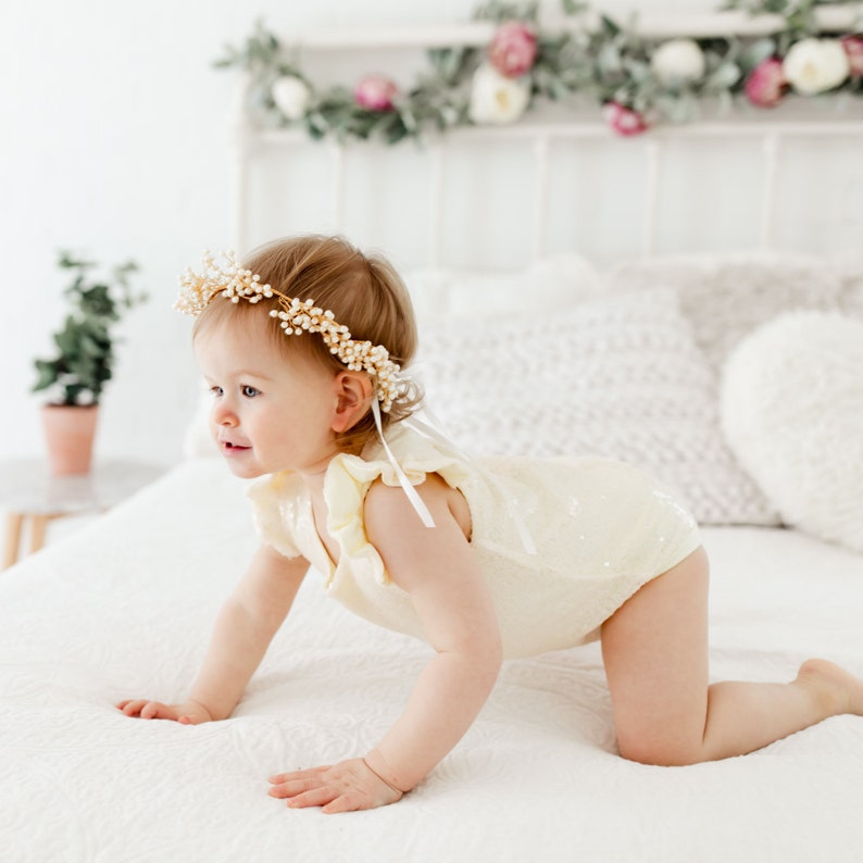 Puede incluir: Una ni&ntilde;a beb&eacute; con un body blanco y amarillo con lentejuelas y una corona de flores de perlas y oro. La beb&eacute; est&aacute; gateando en una colcha blanca.