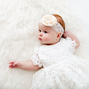 May include: A baby girl wearing a white lace dress and a headband with a flower on it. The baby is lying on a white fluffy blanket.