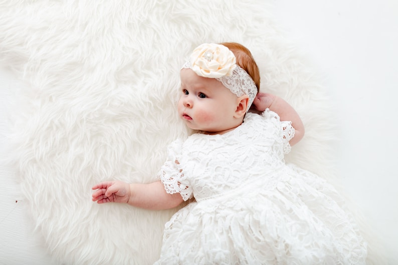 May include: A baby girl wearing a white lace dress and a headband with a flower on it. The baby is lying on a white fluffy blanket.