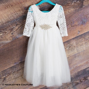 Puede incluir: Vestido blanco de tul con mangas largas de encaje y un cintur&oacute;n de pedrer&iacute;a. El vestido tiene un escote redondo y una falda completa. &copy; Nicolette's Couture