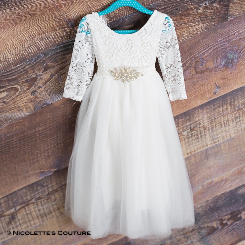 Puede incluir: Vestido blanco de tul con mangas largas de encaje y un cintur&oacute;n de pedrer&iacute;a. El vestido tiene un escote redondo y una falda completa. &copy; Nicolette's Couture