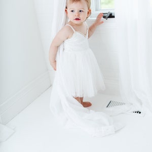 Puede incluir: Una ni&ntilde;a peque&ntilde;a con un vestido blanco de tul con detalles de encaje se encuentra en una habitaci&oacute;n blanca, asom&aacute;ndose desde detr&aacute;s de una cortina blanca.