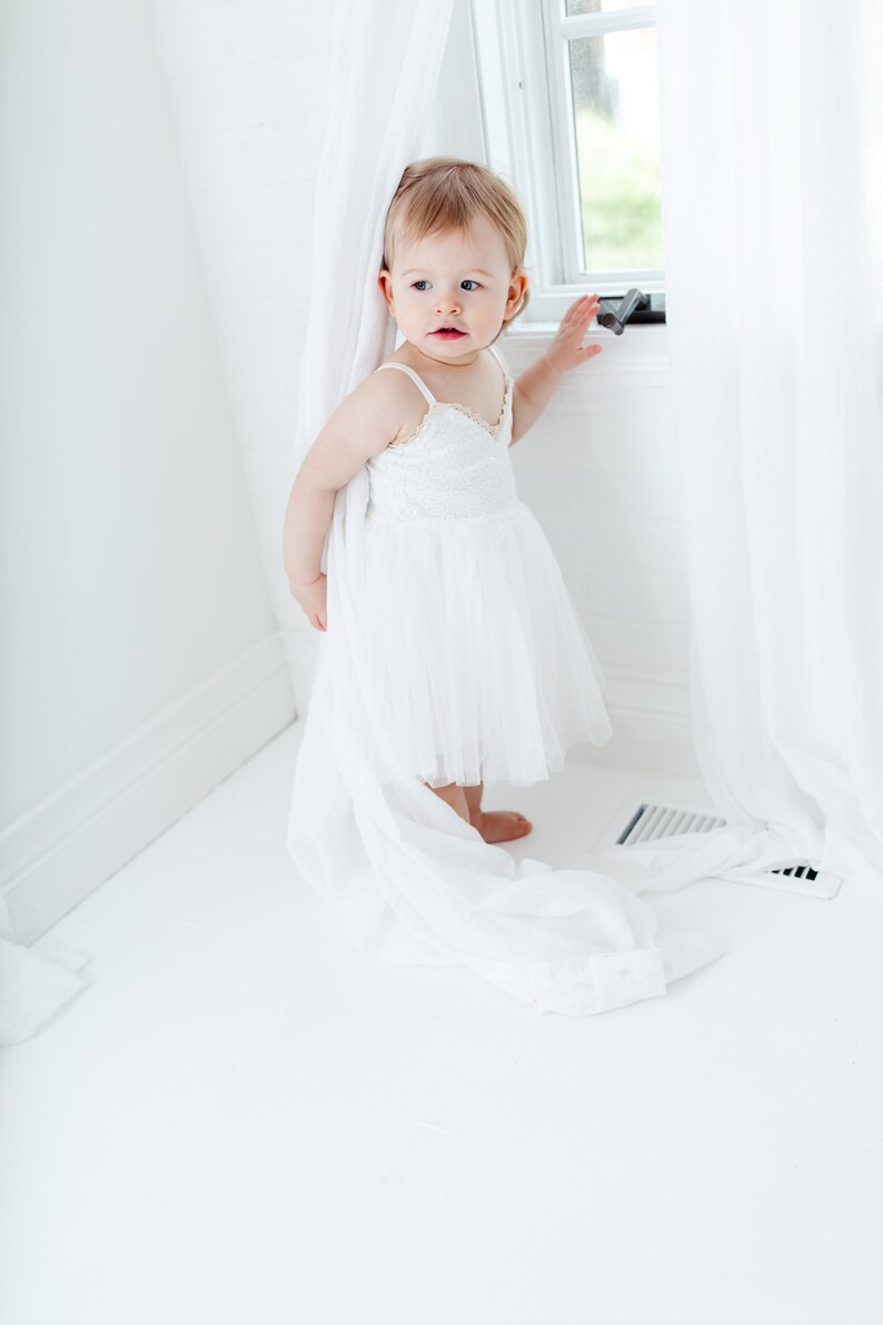 Puede incluir: Una ni&ntilde;a peque&ntilde;a con un vestido blanco de tul con detalles de encaje se encuentra en una habitaci&oacute;n blanca, asom&aacute;ndose desde detr&aacute;s de una cortina blanca.