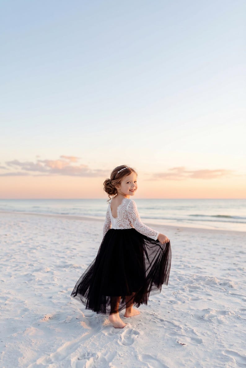 May include: A long-sleeved dress featuring a white lace bodice with a scoop neckline and a full-length black tulle skirt. The garment has a fitted lace top that transitions into a voluminous, layered black skirt that reaches the ankles.