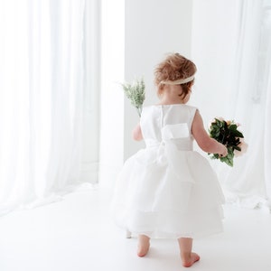Puede incluir: Una ni&ntilde;a peque&ntilde;a con un vestido blanco sostiene un ramo de flores rosas y verdes. Est&aacute; de pie sobre una superficie blanca con una cortina blanca en el fondo.