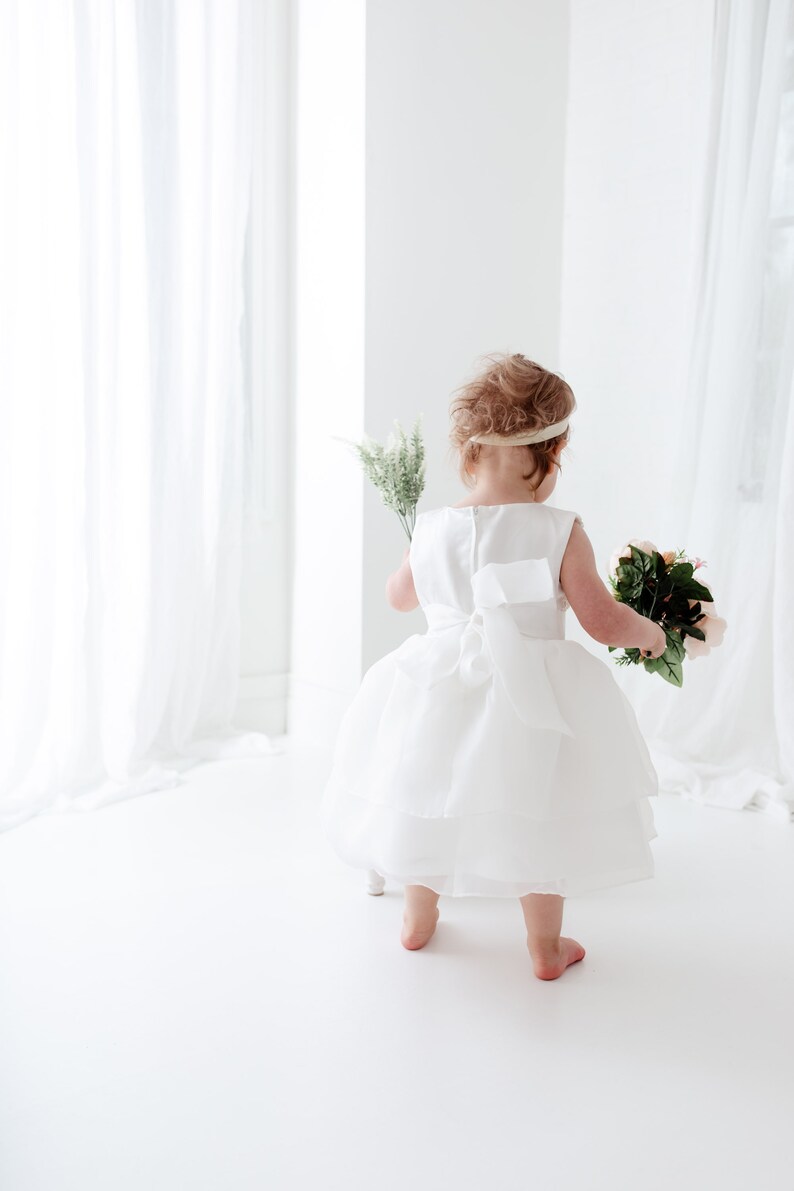 Puede incluir: Una ni&ntilde;a peque&ntilde;a con un vestido blanco sostiene un ramo de flores rosas y verdes. Est&aacute; de pie sobre una superficie blanca con una cortina blanca en el fondo.