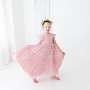 May include: A young girl wearing a pink, tiered, pleated dress with lace detailing and a floral crown. The dress has short, ruffled sleeves and a flowy skirt.