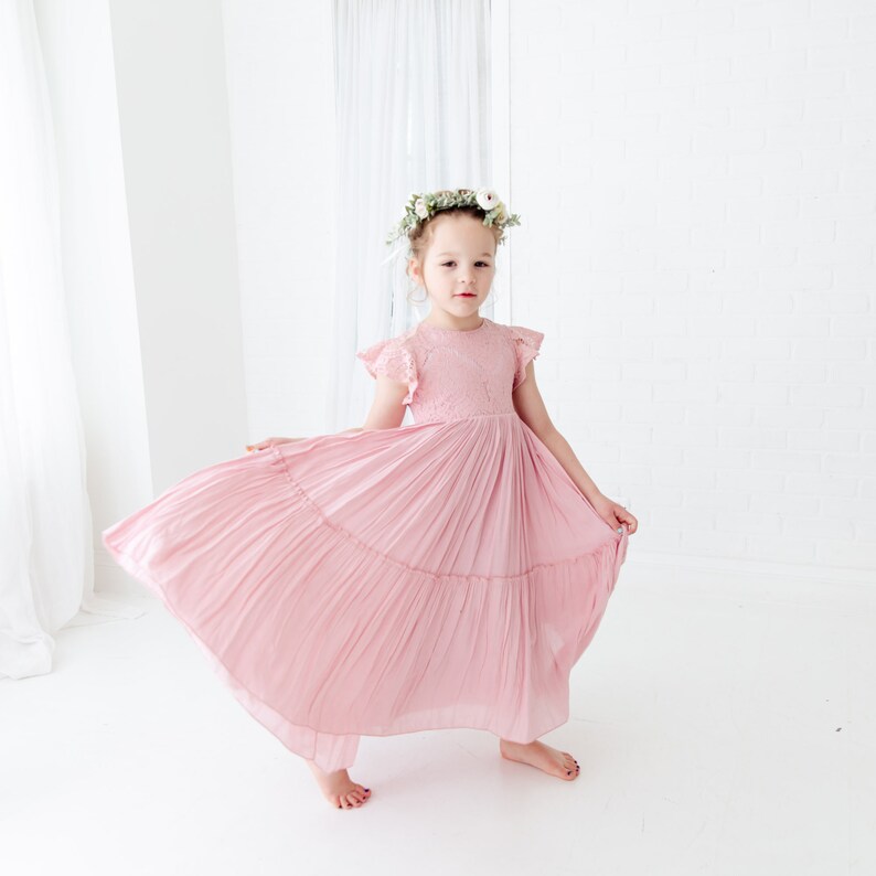May include: A young girl wearing a pink, tiered, pleated dress with lace detailing and a floral crown. The dress has short, ruffled sleeves and a flowy skirt.