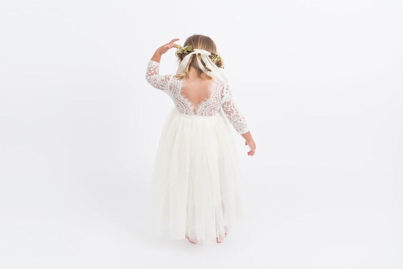 Pu&ograve; includere: Un abito bianco di tulle con un corpetto in pizzo e maniche lunghe. L'abito ha una schiena bassa con un design in pizzo. La ragazza indossa una corona di fiori.