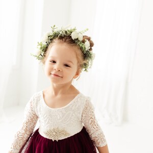 May include: A young girl wearing a white lace dress with a burgundy tulle skirt and a flower crown. She is smiling at the camera.