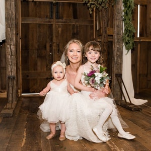 May include: A bride in a white lace wedding dress poses with two flower girls in white dresses. The bride is holding the older flower girl, who is holding a bouquet of purple and white flowers. The younger flower girl is standing in front of the bride. The three are in a rustic barn setting.