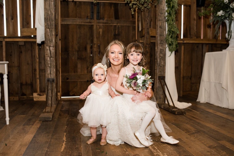 May include: A bride in a white lace wedding dress poses with two flower girls in white dresses. The bride is holding the older flower girl, who is holding a bouquet of purple and white flowers. The younger flower girl is standing in front of the bride. The three are in a rustic barn setting.