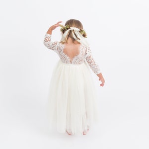 May include: A floor-length white dress with three-quarter length lace sleeves and a lace bodice featuring a deep scoop back. The skirt is made of layered tulle fabric. A floral crown with long white ribbons is worn on the head.