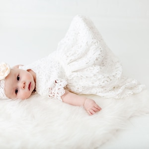 May include: A baby wearing a white lace dress and a headband with a flower on it, lying on a white fluffy rug.