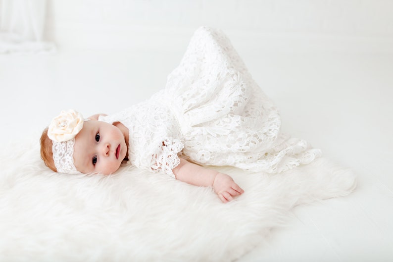 May include: A baby wearing a white lace dress and a headband with a flower on it, lying on a white fluffy rug.