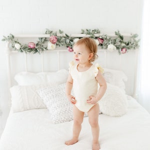 Puede incluir: Un ni&ntilde;o peque&ntilde;o con un body blanco con volantes se para en una cama blanca con una cabecera blanca decorada con una guirnalda de vegetaci&oacute;n y flores rosas.