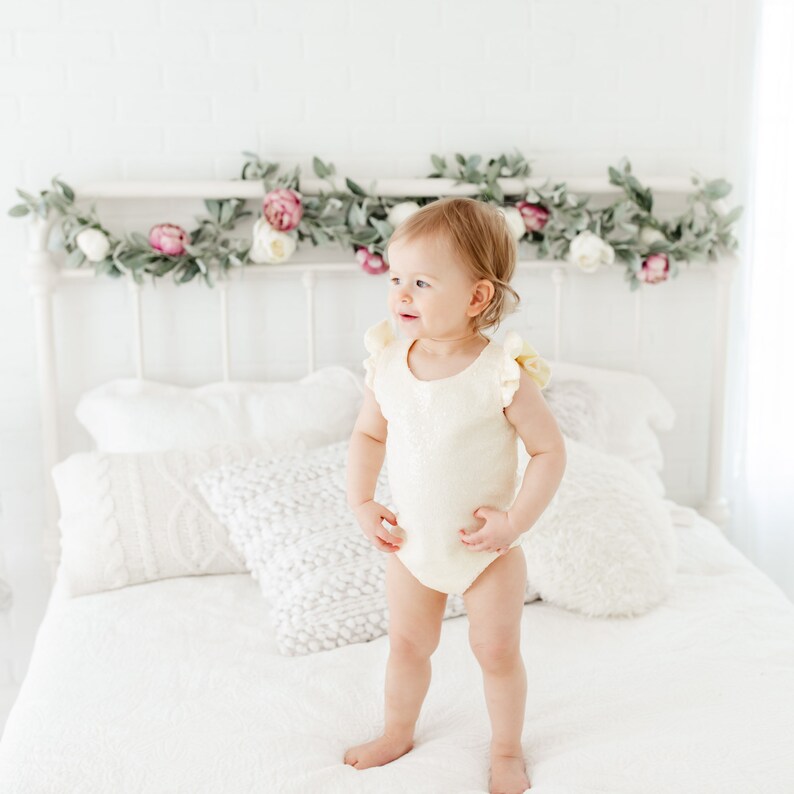 Puede incluir: Un ni&ntilde;o peque&ntilde;o con un body blanco con volantes se para en una cama blanca con una cabecera blanca decorada con una guirnalda de vegetaci&oacute;n y flores rosas.