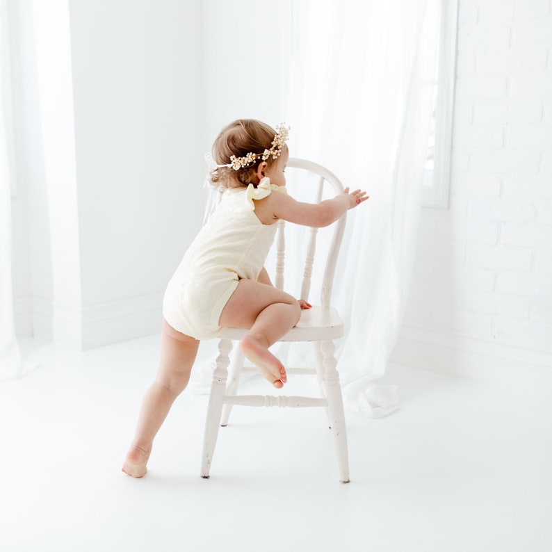 Puede incluir: Una ni&ntilde;a peque&ntilde;a con un vestido blanco y una corona de flores est&aacute; subiendo a una silla de madera blanca. La ni&ntilde;a est&aacute; de espaldas a la c&aacute;mara y est&aacute; alcanzando algo fuera de cuadro.