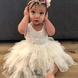 Op de afbeelding: Een babymeisje draagt een witte tule jurk met een bloemenhoofdband. De jurk heeft een kantbovenstuk en een gelaagde tule rok. De hoofdband is versierd met roze en witte bloemen.