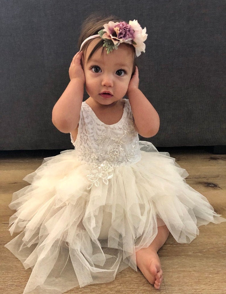 Op de afbeelding: Een babymeisje draagt een witte tule jurk met een bloemenhoofdband. De jurk heeft een kantbovenstuk en een gelaagde tule rok. De hoofdband is versierd met roze en witte bloemen.