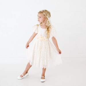 May include: A cream-colored lace dress featuring short flutter sleeves, a round neckline, and a waist adorned with three large fabric flowers. The skirt falls to the knee with a scalloped lace hem, paired with simple white flat shoes.