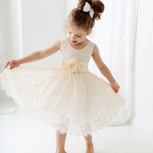 May include: A young girl wearing a white lace dress with a flower embellishment on the waist. She is standing on a white surface and has her hair pulled back with a white bow.
