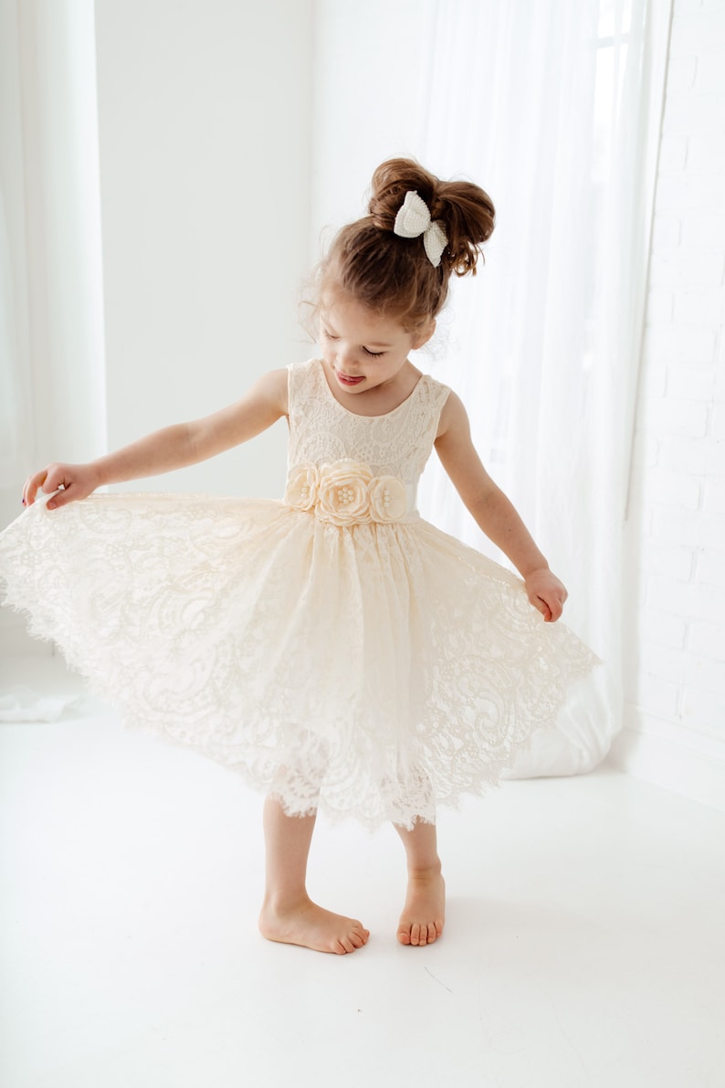 May include: A young girl wearing a white lace dress with a flower embellishment on the waist. She is standing on a white surface and has her hair pulled back with a white bow.