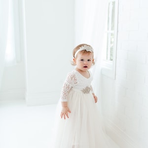 Pu&ograve; includere: Una bambina che indossa un abito bianco di pizzo con gonna di tulle e una fascia scintillante. &Egrave; in piedi in una stanza bianca con una finestra sullo sfondo.