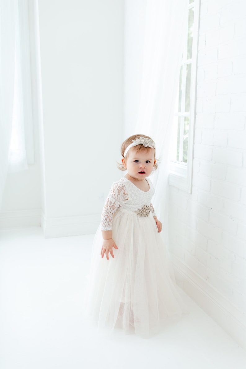 Pu&ograve; includere: Una bambina che indossa un abito bianco di pizzo con gonna di tulle e una fascia scintillante. &Egrave; in piedi in una stanza bianca con una finestra sullo sfondo.