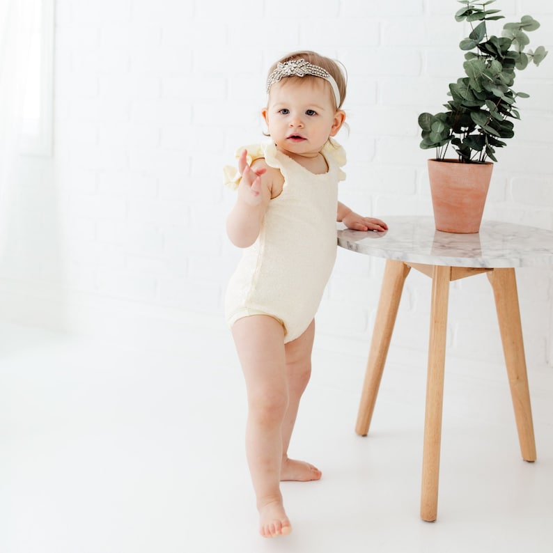 Puede incluir: Una ni&ntilde;a peque&ntilde;a con un mono amarillo con volantes y una diadema brillante se encuentra junto a una mesa peque&ntilde;a, redonda, con tapa de m&aacute;rmol y patas de madera.