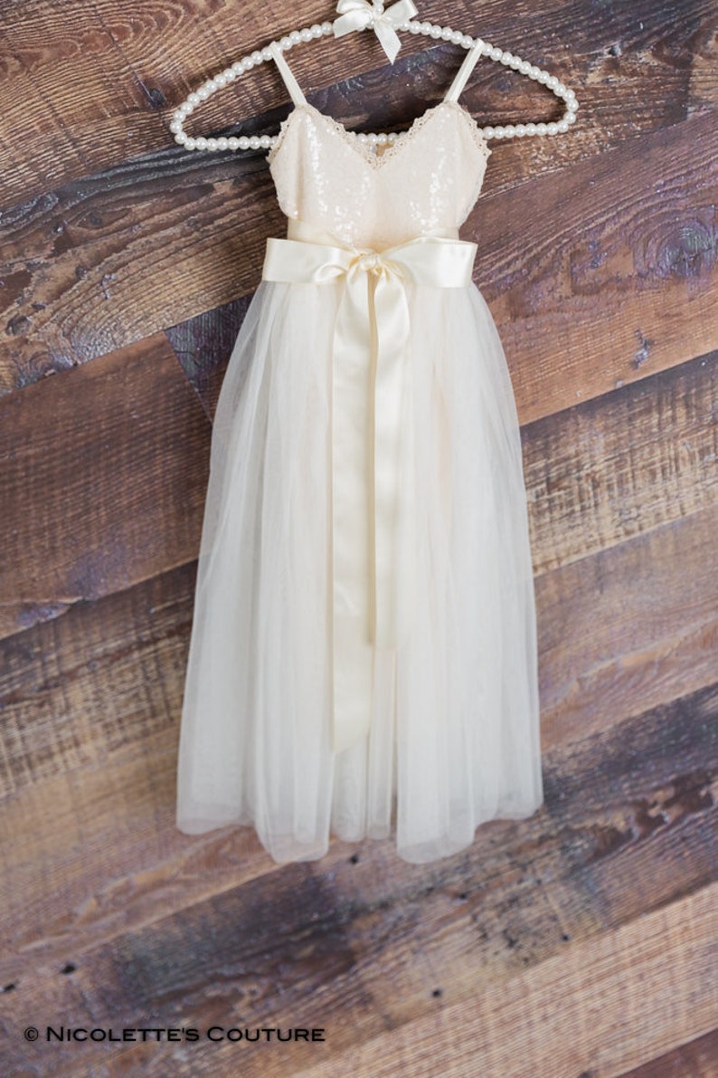 May include: A white tulle dress with a sequined bodice and a satin sash. The dress is hanging on a pearl-beaded hanger.