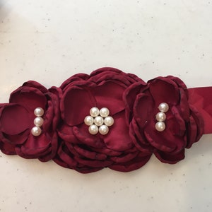 May include: A burgundy satin sash with three large fabric flowers, each adorned with pearl-like embellishments.